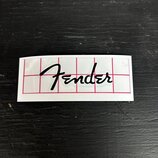 Bílý obdélníkový štítek s černým nápisem Fender v kurzívě. Povrch je proříznut červenou mřížkou s tenkými čarami. Štítek je umístěn na tmavém, textur…