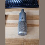 Stříbrný kondenzátorový mikrofon s kovovou síťkou v dolní části. Na těle je černý nápis Studio Projects B1 a logo ve tvaru trojúhelníku. Povrch mikro…
