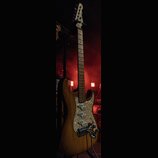 Elektrická kytara s přírodním dřevěným povrchem korpusu a hmatníkem ze světlého dřeva. Kytara má perleťový pickguard a tři snímače typu single-coil.…