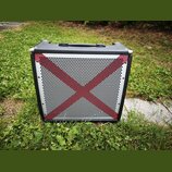 Černý kytarový kombozesilovač s kovovými rohy a rukojetí. Přední mřížka je stříbrná, perforovaná a obsahuje červený kříž. Na přístroji nejsou vidět ž…