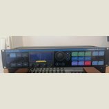 Černý rackový procesor s nápisy TC-HELICON vlevo nahoře a VOICELIVE RACK vpravo nahoře. Zařízení má centrální displej, který je překryt ochrannou fól…
