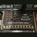 Šedý analogový drum synthesizer značky jomox, model XBASE 999. Zařízení má mnoho ovládacích knoflíků, posuvníků a bílá tlačítka v dolní části s popis…
