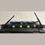 Černý rackový bezdrátový přijímač s dvěma anténami po stranách. Na předním panelu jsou čtyři ovládací prvky označené žlutými samolepkami s čísly 1, 2…