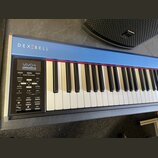 Digitální piano v modré barvě s bílými a černými klávesami. Na horní liště je bílý nápis DEXIBELL a v ovládacím panelu nápisy VIVO S1, Grand Piano Mo…