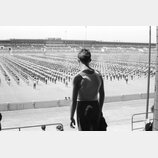 Na fotografii není žádný hudební nástroj ani příslušenství. Snímek zobrazuje muže v tílku zády k fotografu, který se dívá na rozsáhlý shromážděný dav…