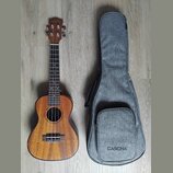 Dřevěná ukulele v přirozené hnědé barvě s černým hmatníkem a čtyřmi strunami. Na hlavě nástroje i na spodní části šedého textilního obalu je uveden n…