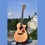 Akustická kytara s cutawayem v přirozené dřevěné barvě, umístěná na černém stojanu. Na hlavě hmatníku je bílý nápis Taylor. Kytara má hnědý hmatník,…
