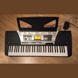 Černý digitální klavír s bílými a černými klávesami a přiloženým napájecím adaptérem. Na ovládacím panelu je nápis YAMAHA a model PSR-350, dále označ…