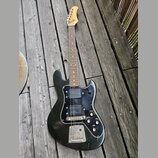 Černá elektrická kytara s dřevěným hmatníkem a hlavou, položená na dřevěné terase. Nástroj má černý pickguard a dva snímače typu humbucker. Povrch tě…