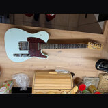 Světle modrá elektrická kytara typu Telecaster s červeně hnědým vzorovaným pickguardem a hnedým hmatníkem. Na hlavě kytary je vidět nápis Fender TELE…