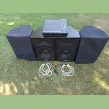 Sada zvukové techniky složená z mixážního pultu, dvou černých reproduktorů a dvou černých krytů z látky. Na přední straně obou reproduktorů je umístě…
