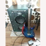 Modrá elektrická kytara s tmavým hnedým hmatníkem a červeným popruhem. Kytara stojí před černým kytarovým kombem, které má v horní části nápis "ROCK…