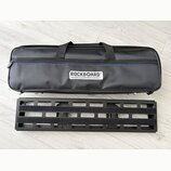 Černý textilní softcase s rukojetmi a modrým detailem u zipu. Na přední straně je bílý štítek s nápisem ROCKBOARD® by WARWICK. Pod pouzdrem leží čern…