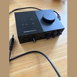 Černý audio rozhraní s nápisem KOMPLETE AUDIO 1. Na horní straně je velký ovládací knoflík, nápisy USB, 48 V a OUT 1, dále indikátory IN 1 a IN 2. Př…