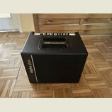 Černý kombozesilovač s texturovaným povrchem a integrovanými madly. Na horní části je nápis AER The Acoustic People a na boční straně vertikální bílý…