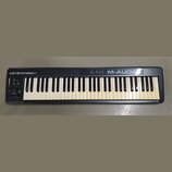 Černá klaviatura s bílými a černými klávesami. Na horní liště je vlevo nápis KEYSTATION61 a vpravo M-AUDIO. V levé části jsou umístěny posuvníky, dva…