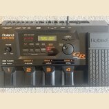 Černý kytarový syntetizátor Roland GR-33 Guitar Synthesizer. Zařízení má digitální displej s nápisem Roland GR-33 Guitar Synth, několik ovládacích tl…