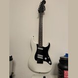 Bílá elektrická kytara s černým hmatníkem a černým pickguardem. Na hlavě je bílý nápis Squier STRATOCASTER. Nástroj má tři snímače, jeden ovládací kn…