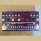 Bílo-fialový analogový syntetizátor s nápisy TD-3 Computer Controlled a Analog Bass Line Synthesizer. Na přístroji je viditelné logo behringer a řada…