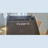 Černý osobní monitor s kovovou mřížkou, na které je bílý nápis Roland. Horní ovládací panel obsahuje vstupy INPUT (V-Drums, LINE), ovladače VOLUME (V…