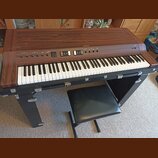 Digitální piano v dřevěném korpusu s hnědým povrchem, vybavené klávesnicí a černou stoličkou. Na horním panelu je nápis YAMAHA a řada ovládacích prvk…