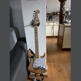 Elektrická kytara s přírodním dřevěným povrchem těla a světlou hmatníkovou deskou. Na hlavě je viditelné logo "SG". Kytara má bílý pickguard s textur…