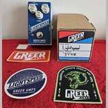 Na fotografii je efekt pedál stříbrné barvy s nápisy LIGHTSPEED, ORGANIC OVERDRIVE a GREER AMPS PRODUCT OF ATHENS, GEORGIA. Vedle pedálu je hnědá kar…