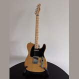 Elektrická kytara typu Telecaster v přírodní dřevěné barvě s černým pickguardem. Na hlavě hmatníku je vidět nápis Squier. Nástroj je umístěn na černé…