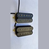 Dvě černé elektrické kytarové snímače typu humbucker s kovovými zlatými rámečky. Každý snímač má dvě řady kulatých pólů. Povrch plastových krytů je h…
