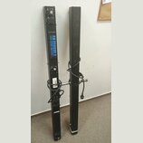 Dvě černé, úzké a vysoké lineární reproduktorové sloupy z kovového materiálu. Levý sloup obsahuje panel s modrými prvky, několika konektory a přepína…