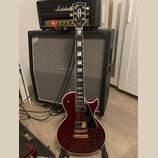 Červená elektrická kytara typu Les Paul s bílým bindingem a zlatými potenciometry a hardwarem. Na hlavě hmatníku je nápis Gibson a logo, na hmatníku…