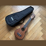 Hnědý dřevěný ukulele s černým hmatníkem a černým mostkem. Na hlavě nástroje je bílý nápis CASOCHA. Vedle nástroje leží černý textilní obal s bílým n…