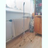 Kovový mikrofonní stojan s trojnožkou a teleskopickým horním ramenem. Povrch je stříbrný, v místě spojů jsou vidět utahovací šrouby. Na stojanu nejso…