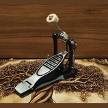 Fotografie zobrazuje jednopedálový basový pedál s černým základem a stříbrným, kovovým šlapadlem. Mechanismus obsahuje řetězový pohon a beater s bílý…