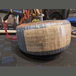 Fotografie znázorňuje elektrický transformátor s modrým lakovaným jádrem, omotaný průhlednou fólií. Na boku je nalepený papírový štítek s ručně psaný…