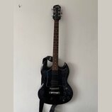 Černá elektrická kytara s tvarem SG a tmavým hnedým hmatníkem. Na hlavě je bílý nápis Epiphone a na černém popruhu nápis Fender. Kytara má dva humbuc…