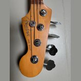 Detail hlavy basové kytary z přírodního dřeva. Na dřevě je černý nápis Fender, pod ním menší text MADE IN U.S.A. a vedle něj JAZZ BASS®. Nástroj má č…