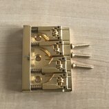 Zlatý kovový kobylkový mechanismus s třemi saddlesy a třemi přiloženými šrouby. Povrch je lesklý, bez viditelných značek, log nebo nápisů. Předmět vy…