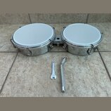 Dvojice bongos s bílými membránami a chromovaným kovovým korpusem. Nástroje jsou spojeny kovovou spojkou. Před nimi leží dva kovové nástroje pro ladě…