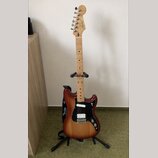 Elektrická kytara s tělem v červenohnědém sunburst provedení a světlým javorovým hmatníkem. Na hlavě je viditelné logo Fender a na popruhu nápis Fend…