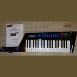 Černý MIDI kontrolér s klávesnicí a modrým podsvícením displeje a indikátorů. Na přístroji je bílý nápis ALESIS VORTEX WIRELESS 2. Vedle nástroje lež…