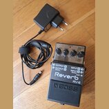 Na dřevěné podlaze leží efekt pedál Boss RV-6 Reverb v černé barvě s kovovým horním panelem a černý síťový adaptér. Na pedálu jsou nápisy „Reverb“, „…