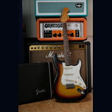 Elektrická kytara typu Stratocaster s barevným přechodem od žluté přes oranžovou k hnědé a bílým pickguardem. Na hlavě krku je viditelné logo Fender.…