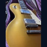 Detail elektrické kytary typu Les Paul v zlatavé barvě, která leží v otevřeném kufru s fialovou výstelkou. Na těle jsou dva snímače v bílých rámečcíc…