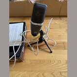 Stříbrný mikrofon s černou mřížkou umístěný na černém tripodním stativu. Na těle mikrofonu jsou nápisy MONITOR, 22040955 a ABx555. Vedle mikrofonu je…