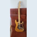 Elektrická kytara typu Stratocaster s přírodním dřevěným povrchem těla a hmatníkem. Tělo má tmavě červený želvový pickguard a tři bílé snímače. Na hl…