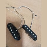 Dvě černé elektrické snímače typu humbucker s pěti viditelnými kovovými póly každý. Snímače mají připojené bílé a černé vodiče s odhalenými konci. Na…