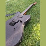 Černá elektrická kytara s matným povrchem a viditnou strukturou dřeva, položená na trávě. Nástroj má dva humbuckery, jeden ovládací knoflík a černý m…