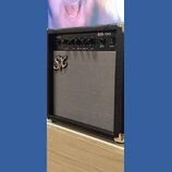 Kytarový kombo zesilovač v černém pouzdře s šedou textilní maskou reproduktoru. Na předním panelu jsou čtyři ovládací knoflíky s popisky VOLUME, HIGH…