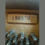 Detailní záběr na část hudebního nástroje se zlatým povrchem. Na obdélníkovém štítku je černý nápis 199576. Ve spodní části jsou vidět kovové kolíky…